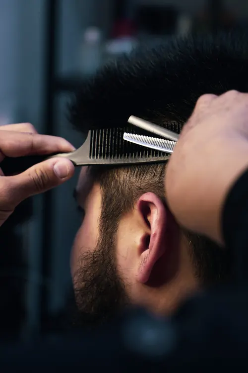 Barber podnosi grzebieniem włosy klienta i przerzedza je nożyczkami